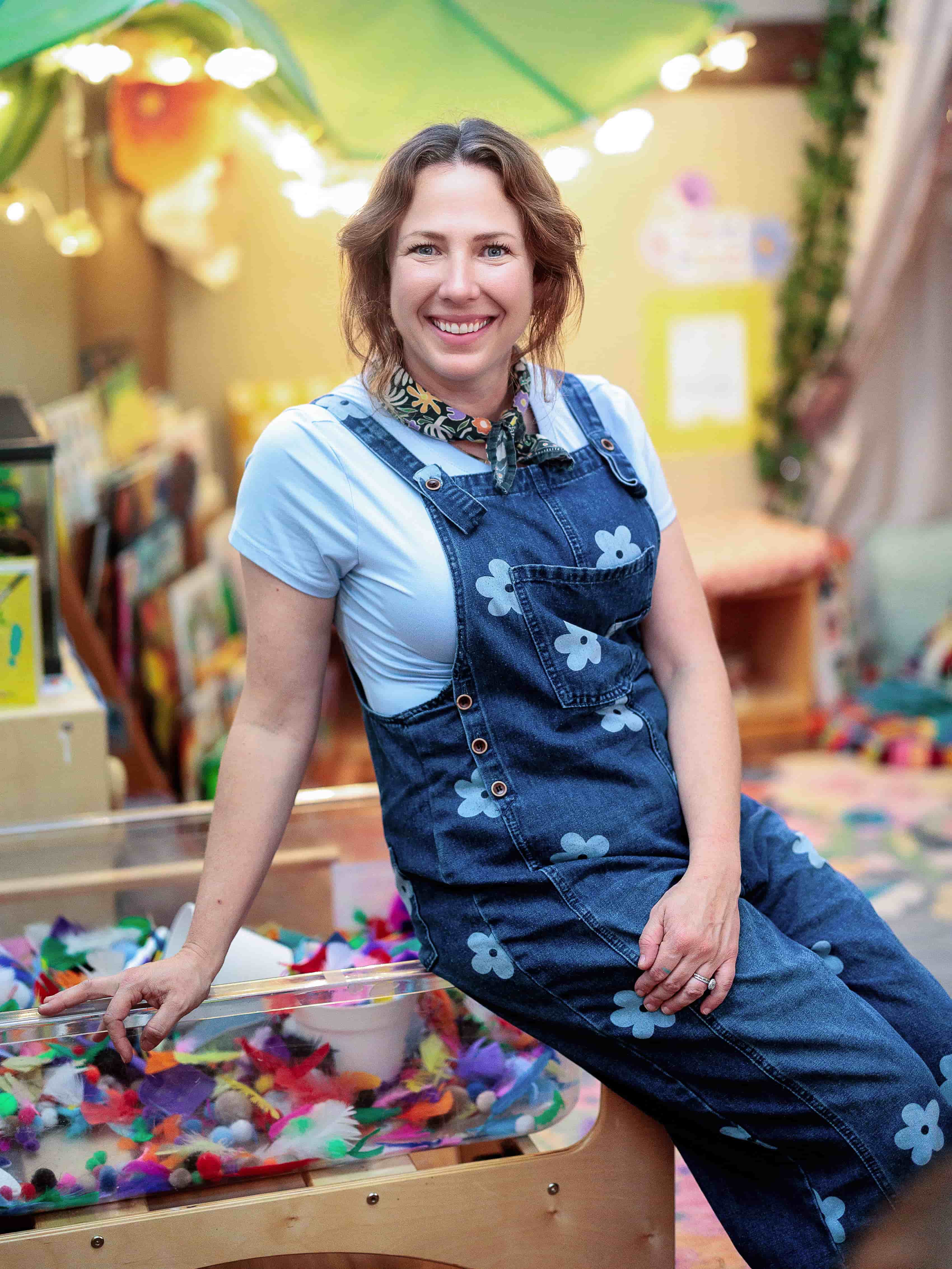 Sarah Anne Stephens smiling in the Storybook Studio classroom beside a colorful sensory table